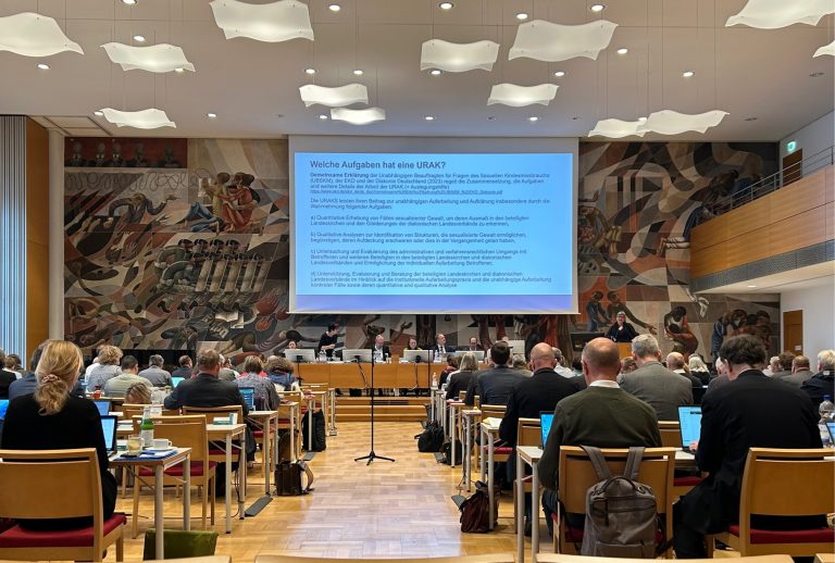 Die URAK-Vorsitzende Heide Glaesmer spricht vor den Synodalen an einem Rednerpult. Zu sehen ist ihre Präsentation zur Arbeit der Aufarbeitungskommission. In der Mitte sitzen mehrere Personen an einem Tisch (Podium). An der Wand hinter ihr und der Leinwand ist ein Flächendeckendes ein modernes Kunstwerk zu sehen.