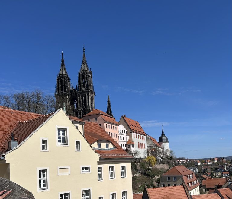 Blick auf die Stadt Meißen mit ihrem Dom mit einem hellen Haus im Vordergrund und weiteren Häusern mit roten Dächern im Hintergrund.