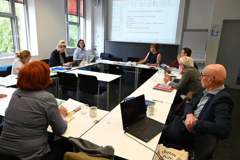 Eine Gruppe von Männer und Frauen sitzen an Tischen vor einer Leinwand mit einer Präsentation, im Vordergrund ist ein Laptop zu sehen.
